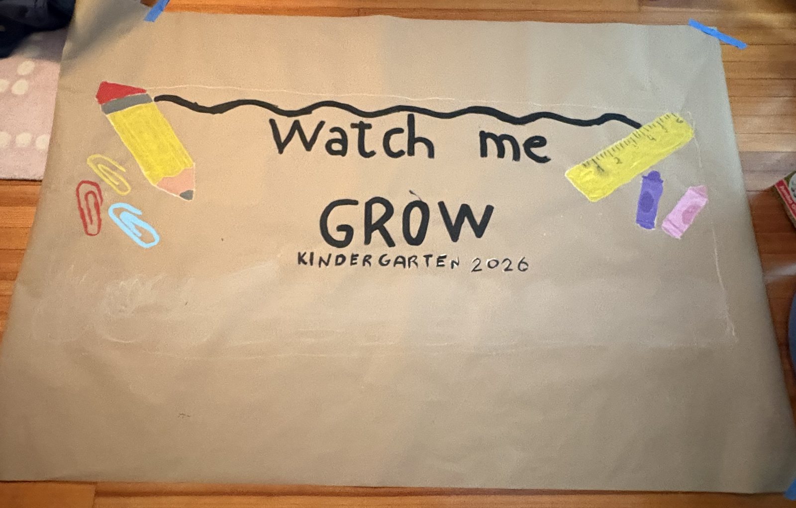 Hand-painted kraft paper banner reading Watch me Grow, Kindergarten 2026, with a painted pencil, ruler, crayons and paper clips
