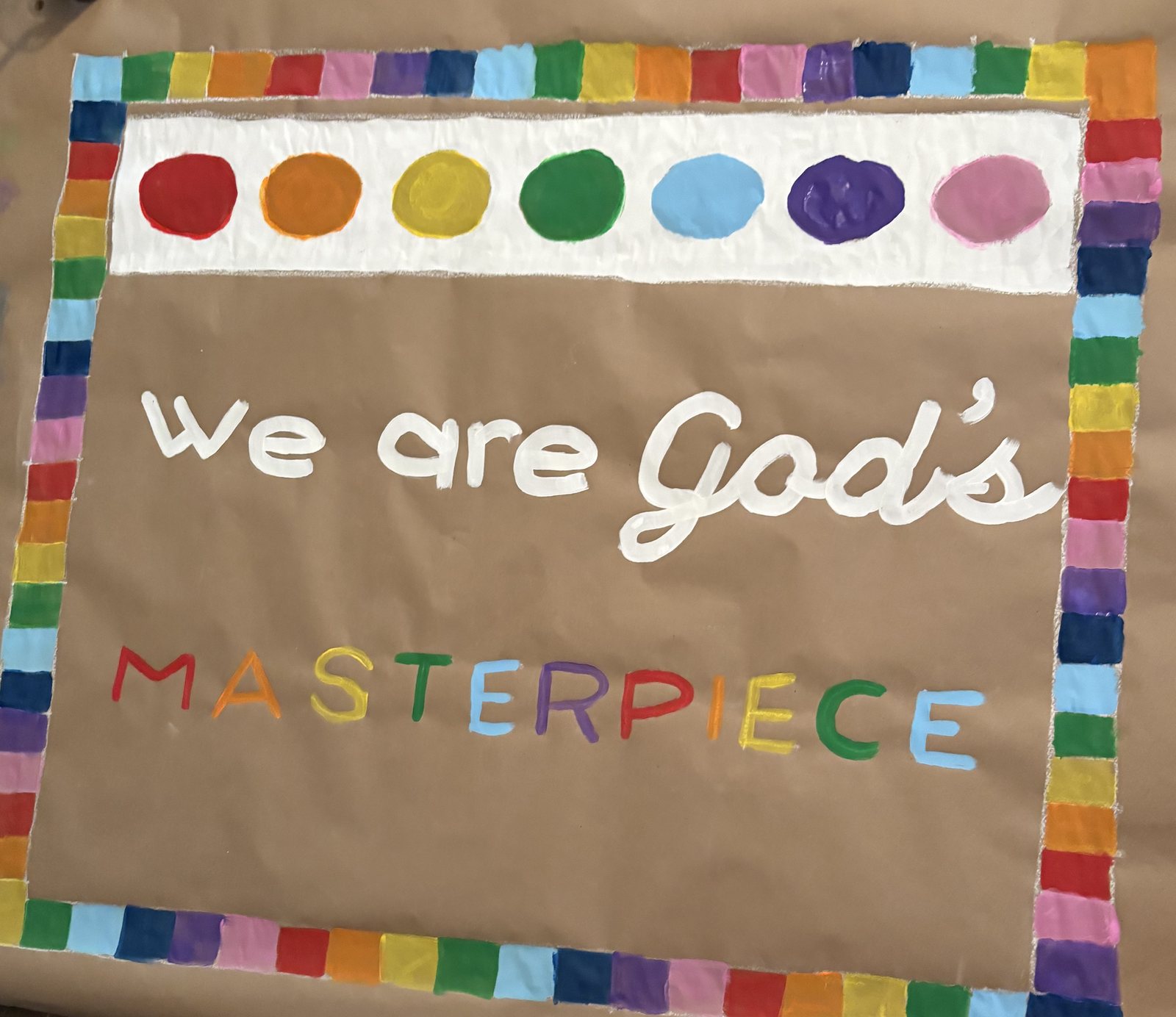 Hand-painted banner reading We are God's Masterpiece with a rainbow block border and a row of painted paint dots